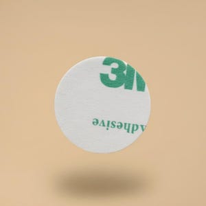 May include: A white, circular adhesive pad with green "3M" branding and the word "adhesive" printed on it. The pad is floating against a light brown background.