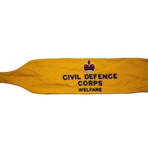 Könnte beinhalten: Senfgelber Civil Defence Corps-Wohlfahrtsgürtel. Der Gürtel hat einen Schnallenverschluss und zeigt das Logo und den Text des Civil Defence Corps in Schwarz. Der Gürtel hat eine lange, schmale Form mit einem abgerundeten Ende.