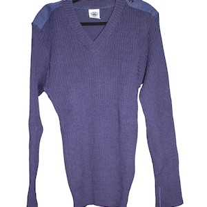 May include: A purple V-neck jumper with long sleeves. The jumper features a ribbed knit texture and shoulder patches. A small label is visible at the neckline. The garment is a solid colour and has a classic design.