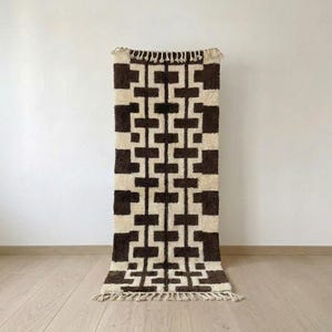 Peut inclure: Un tapis rectangulaire à poils longs présentant un motif géométrique répétitif en brun foncé et crème. Le design se compose de formes en blocs imbriquées disposées verticalement, avec des franges de couleur crème aux deux extrémités.