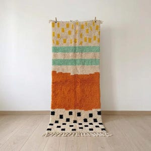 Peut inclure: Un tapis rectangulaire à poils longs sur une base de couleur crème. Le motif se compose de bandes horizontales avec des tirets jaunes, deux rayures vert clair, un grand bloc orange uni et une section inférieure ornée de carrés noirs.