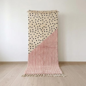 May include: A rectangular area rug featuring a split design. The top section is cream-colored with small black square patterns, while the bottom section is a dusty rose pink with vertical white stripes. The rug has fringe trim on both ends.