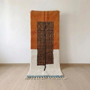 May include: A rectangular area rug featuring a color-blocked design with a burnt orange top section and a cream bottom section. A central dark brown grid pattern runs vertically through the middle, finished with a light blue border and fringe.