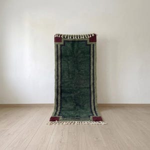 Könnte beinhalten: Ein rechteckiger Teppich mit einem tiefen waldgrünen Mittelfeld, das von dunkelblauen Rändern und bordeauxroten Eckakzenten eingerahmt wird. Der Teppich ist an den oberen und unteren Kanten mit cremefarbenen, geknoteten Fransen versehen.