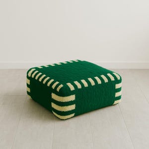 May include: A square, green ottoman with cream-colored, striped trim. The ottoman is made of a soft, textured material and sits on a light-colored tile floor. The design is modern and minimalist.
