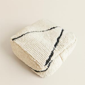 Könnte beinhalten: Quadratischer, cremefarbener Pouf mit einer strukturierten, gerippten Oberfläche und schwarzen, diagonalen Streifen. Der Pouf hat eine Würfelform und besteht wahrscheinlich aus einem weichen, plüschigen Material. Dieses dekorative Akzentstück verleiht jedem Wohnraum einen Hauch von Textur und Stil.