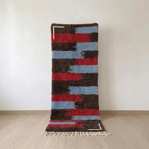 Peut inclure: Un tapis rectangulaire en laine à poils longs présentant un motif à rayures horizontales dans des tons brun foncé, rouge vif et bleu clair. Le design comprend deux petits accents blancs dans les coins et une frange crème sur le bord inférieur.