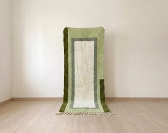 May include: A rectangular area rug featuring a central cream-coloured panel framed by concentric borders in sage green and dark olive green. The rug has a textured pile surface and is finished with short, cream-coloured fringe along the bottom edge.