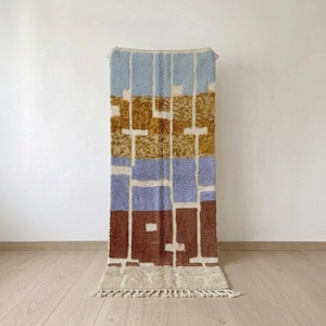 Könnte beinhalten: Ein rechteckiger Teppich mit einem geometrischen Blockmuster in den Farben Hellblau, Senfgelb, Lavendel und Dunkelbraun, getrennt durch dicke cremefarbene Linien. Der Teppich hat einen Hochflor und cremefarbene Fransen an den Enden.