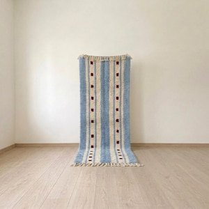 Peut inclure: Un tapis rectangulaire orné de rayures verticales bleu clair et crème. Le motif comprend deux bandes de couleur crème agrémentées d'une série répétitive de petits carrés rouge foncé. Le tapis est terminé par des franges aux deux extrémités.