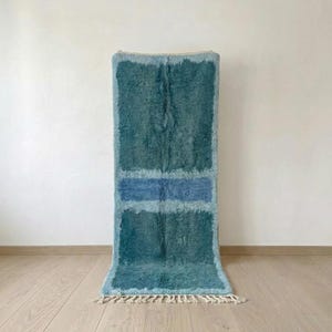 Teal Moroccan wool runner rug with pale blue horizontal bands and fringe on a light studio floor.