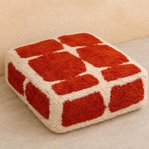 May include: A square, plush ottoman with a brick-like pattern. The design features large, rectangular blocks of vibrant orange against a cream-colored background. The ottoman is a soft furnishing.