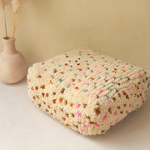 Pode incluir: Uma almofada quadrada texturizada creme com detalhes coloridos. A almofada apresenta um padrão de pequenos tufos em relevo em tons de castanho, laranja, verde-azulado e rosa. Um vaso bege com flores secas está ao fundo.