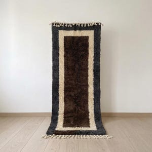 May include: A rectangular area rug featuring a deep brown center panel surrounded by a thick cream-colored border and an outer dark charcoal gray frame. The rug has a shaggy, high-pile texture and includes cream-colored fringe tassels on both ends.