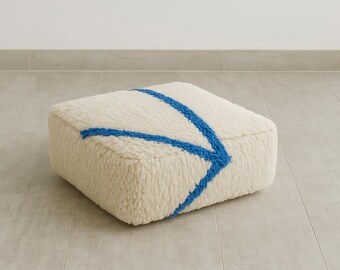 May include: A square floor cushion featuring a thick, cream-colored, high-pile textured fabric. The top surface is decorated with a bold, bright blue yarn-tufted arrow design that points toward the corner of the cushion, creating a distinct contrast.