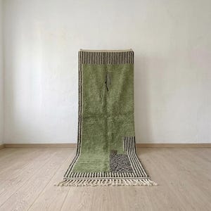 May include: A rectangular wool rug featuring a sage green field with black and white striped borders at the top and bottom. The rug includes a small black and white patterned rectangular accent in the lower right corner and white fringe.