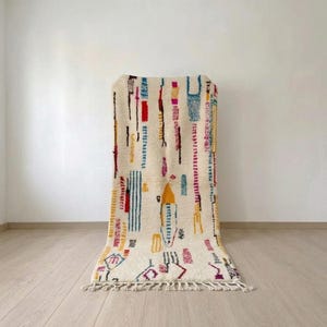 May include: A rectangular off-white area rug featuring a high-pile texture and a series of scattered, colorful geometric patterns in shades of red, yellow, blue, and magenta. The rug is finished with a knotted fringe along the bottom edge.