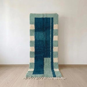 Peut inclure: Un tapis rectangulaire présentant un motif géométrique dans des tons de bleu sarcelle profond, de vert sauge et de crème. Le design comprend des rayures verticales sur les côtés et un grand bloc bleu sarcelle uni au centre. Tapis à poils longs.