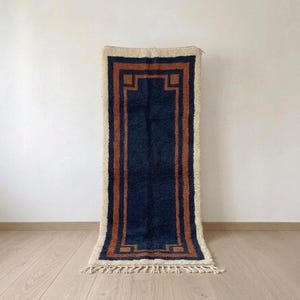 May include: A rectangular area rug featuring a deep navy blue center panel framed by a burnt orange border and a thick cream-colored outer edge. The rug has a shaggy texture and includes decorative fringe detailing along the bottom edge.