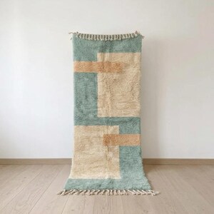May include: A rectangular area rug featuring a high-pile texture with a geometric pattern in sage green and cream. The design includes two large cream-coloured blocks accented by horizontal tan stripes. The rug is finished with cream-coloured fringe.