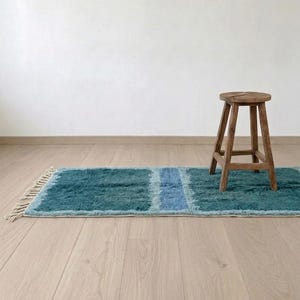 Może przedstawiać: Prostokątny dywan z wzorem w bloki kolor&oacute;w głębokiego błękitu morskiego i jasnego błękitu, o kudłatej fakturze i kremowych frędzlach na jednym końcu. Mały, okrągły, czteronożny drewniany stołek stoi po prawej stronie dywanu.