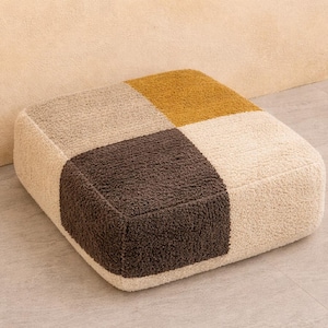 Peut inclure: Un pouf carré avec un motif patchwork. Le dessus est divisé en quatre carrés de couleurs variées : marron, beige, doré et crème. Le pouf a un aspect doux et texturé et est placé sur un sol clair.