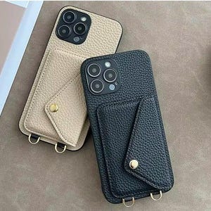 May include: Two leather phone cases, one beige and one black, with attached cardholder pockets. Each case has a gold-colored button closure and a gold-colored metal loop at the bottom for attaching a strap. The cases are designed to fit a smartphone.