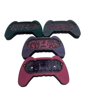 Op de afbeelding: Een verzameling van vijf houten gamecontrollers in verschillende kleuren: donkergroen, donkergrijs, paars en rood. Elke controller heeft een doolhofontwerp in het midden, met kleine goudkleurige accenten.