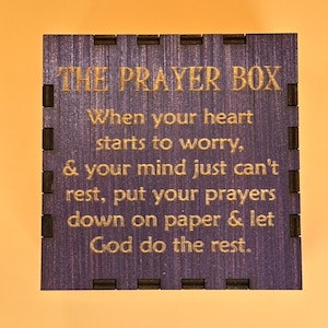 May include: A square wooden box with the text "THE PRAYER BOX" in gold lettering. The box has a dark purple finish with a message about prayer. The box has a decorative edge.