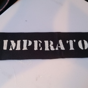 Puede incluir: Parche de tela negro con la palabra "IMPERATOR" impresa en blanco.