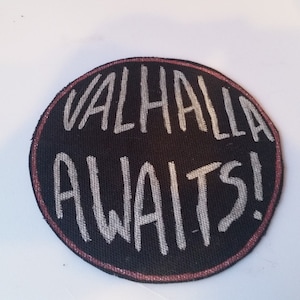 May include: A black round patch with a pink border. The text "VALHALLA AWAITS!" is written in white on the patch.