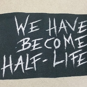 May include: A dark green fabric patch with the words "WE HAVE BECOME HALF-LIFE" in white, handwritten-style lettering. The patch has a slightly frayed edge and a textured appearance.