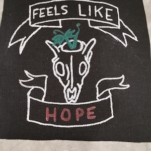 Puede incluir: Parche negro bordado con letras blancas que dicen "Feels Like Hope". Una calavera blanca con una mariposa verde en la parte superior está en el centro del parche.