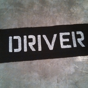 Puede incluir: Parche de tela negro con la palabra "DRIVER" impresa en blanco.
