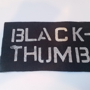 Puede incluir: Parche de tela negro con texto blanco que dice "BLACK-THUMIB".