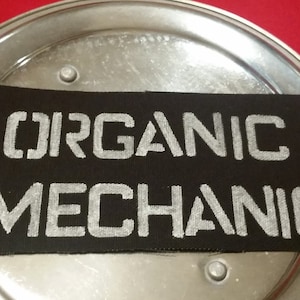 Puede incluir: Un parche rectangular negro con las palabras "ORGANIC MECHANIC" en letras blancas. El parche está sobre una superficie metálica plateada, sobre un fondo rojo.