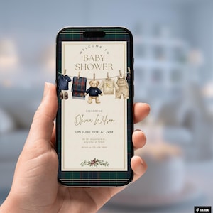 May include: A smartphone displays a baby shower invitation with a plaid border. The invitation features watercolor illustrations of baby clothes and a teddy bear hanging on a clothesline. Text reads "Welcome to Baby Shower" and details for Olivia Wilson.