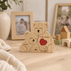 Può includere: Statuetta in legno a forma di famiglia di koala con i nomi "Evelyn", "Alexander" e "Michael" incisi. Un cuore rosso è tra i due koala più grandi. La statuetta è su una superficie di legno, con foto incorniciate sullo sfondo.