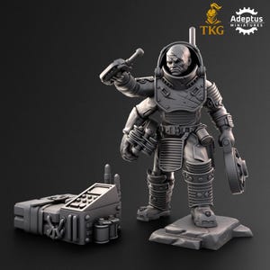 May include: A grey miniature figure of a space marine in a helmet, holding a gun and a circular device. A separate rectangular device with antennas and cylinders is also present. The figure stands on a rocky base. The Adeptus Miniatures logo is visible.