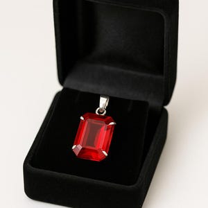 May include: A rectangular, red gemstone pendant with a silver-coloured bail, displayed in an open black velvet jewellery box. The pendant is a vibrant red colour and has a faceted cut. The box is square and has a soft, velvety texture.