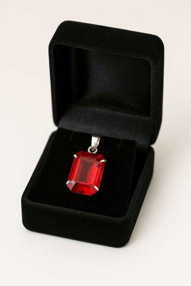 May include: A rectangular, red gemstone pendant with a silver-coloured bail, displayed in an open black velvet jewellery box. The pendant is a vibrant red colour and has a faceted cut. The box is square and has a soft, velvety texture.