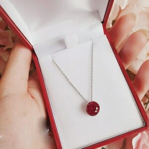 Puede incluir: Un collar de plata con un colgante redondo de piedra preciosa rojo intenso, presentado en una caja de joyería roja con interior blanco. El collar se muestra sobre un fondo suave y borroso.