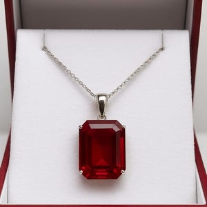 May include: A silver necklace featuring a large, rectangular, deep red gemstone pendant. The pendant is set in a silver bezel and is presented in a red jewellery box with a white interior. The necklace chain is delicate.