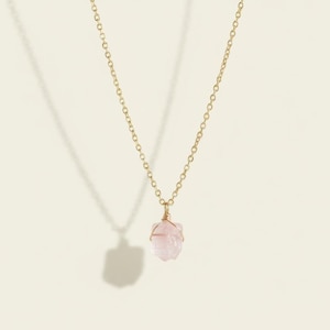 Può includere: Una delicata collana con catena color oro e un piccolo pendente in quarzo rosa chiaro. Il quarzo rosa è avvolto con filo metallico e appeso alla catena. La collana è presentata su uno sfondo color crema.
