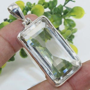 May include: A rectangular pendant featuring a large, clear, faceted gemstone set in a silver metal frame. The pendant has a bail for attaching to a chain. The gemstone has a slight green tint and is multi-faceted.