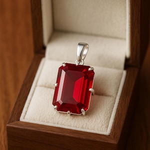 Op de afbeelding: Een rechthoekige, dieprode edelsteen hanger met een zilveren zetting. De hanger wordt getoond in een houten sieradendoos met een crèmekleurig interieur. De edelsteen is ongeveer 2,5 cm lang.