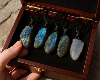 May include: Five raw labradorite pendants on black cords displayed in a wooden box. The stones vary in color, with iridescent blue and green flashes. The box is open, revealing the necklaces against a black velvet lining. The box is a dark brown color.