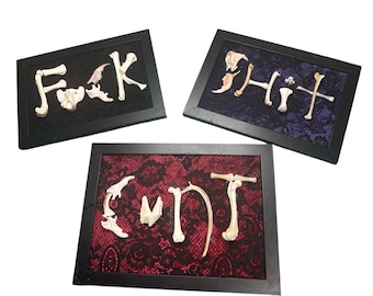 Puede incluir: Tres obras de arte enmarcadas con marcos negros. Cada marco contiene una palabra escrita con fragmentos de hueso. Las palabras son "F*ck", "Sh*t" y "C*nt". Los fondos son de encaje negro, morado y rojo.