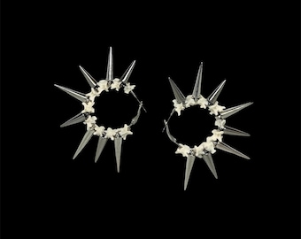May include: A pair of hoop earrings featuring a circular arrangement of small, off-white, bone-like vertebrae segments interspersed with long, pointed, metallic silver-toned spikes extending outward from the centre of each hoop.