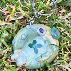 Peut inclure: Un porte-clés en céramique émaillée de forme ronde avec une base vert clair et un motif de trèfle à quatre feuilles vert foncé au centre. La surface est ornée de petits accents circulaires en relief bleus, violets et verts, fixé à un anneau en métal argenté.
